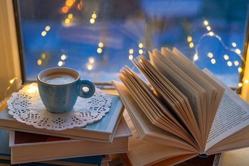 A cozy setting with a cup of coffee and an open book in front of a window lined with fairy lights and it is dusk outside.