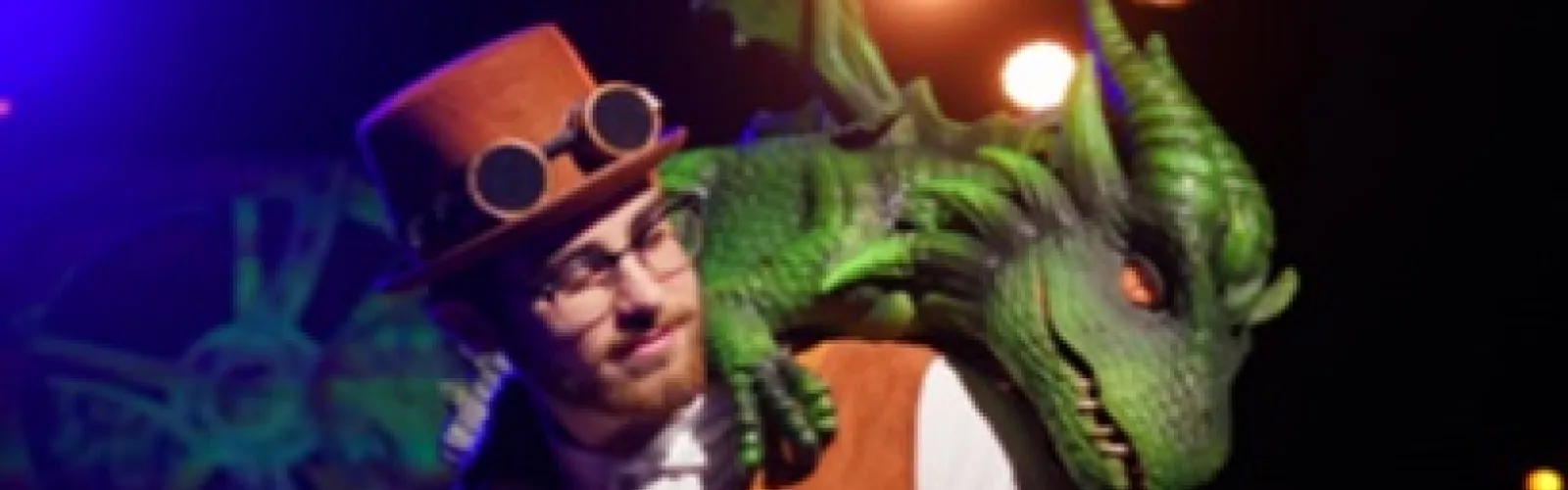 Man in a brown top hat with dark goggles is posing with a green animatronic dragon on his shoulder.